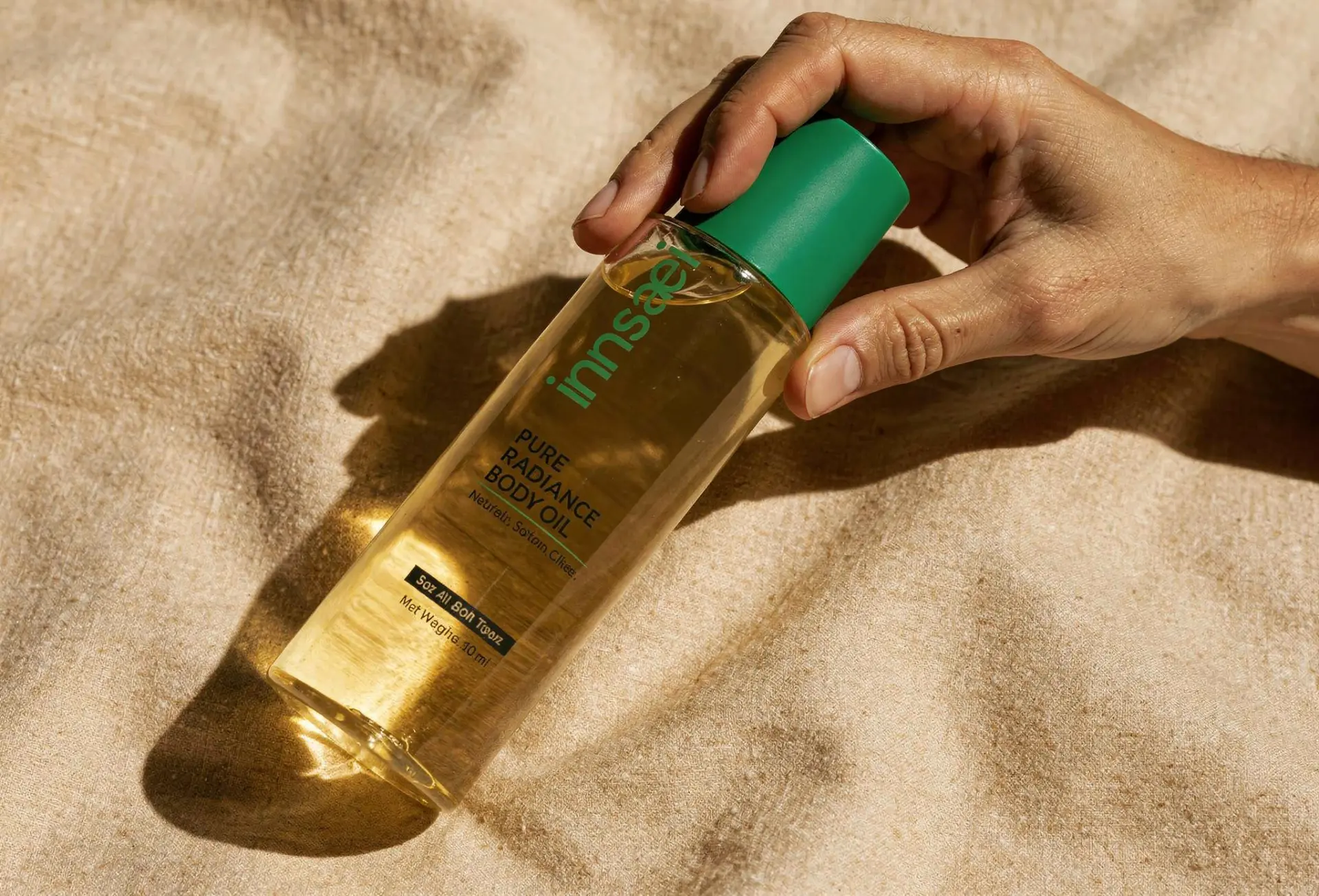 Hand holding a body oil bottle with a green cap on linen in sunlight
